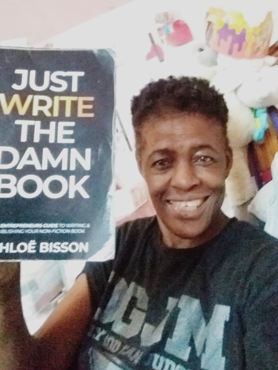 Adedayo smiling and holding up a book about writing, at home.